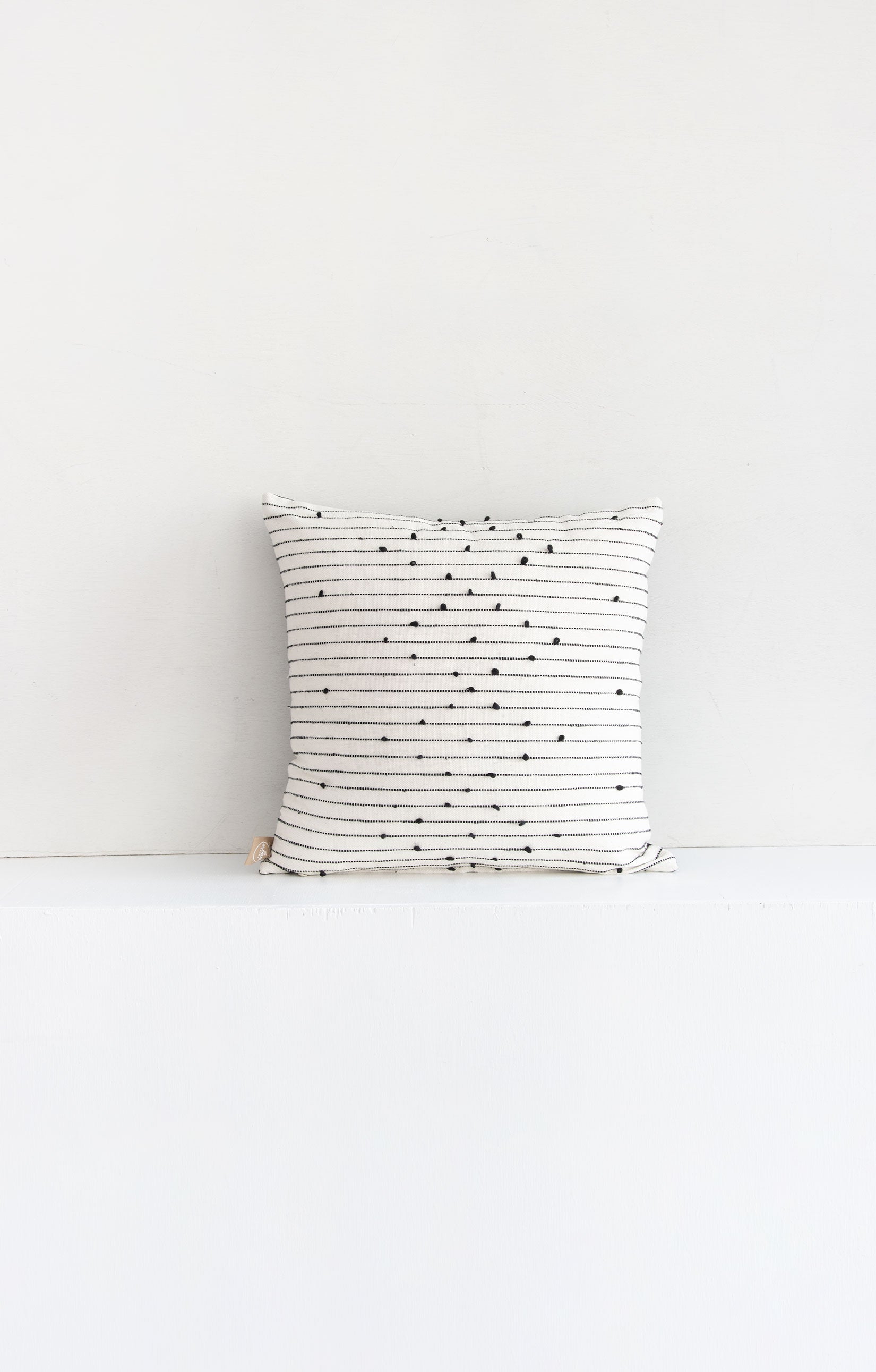 Square white woven throw pillow with small black pom pom accents arranged in a repeating diamond pattern arranged along black lines stretching across the pillow.