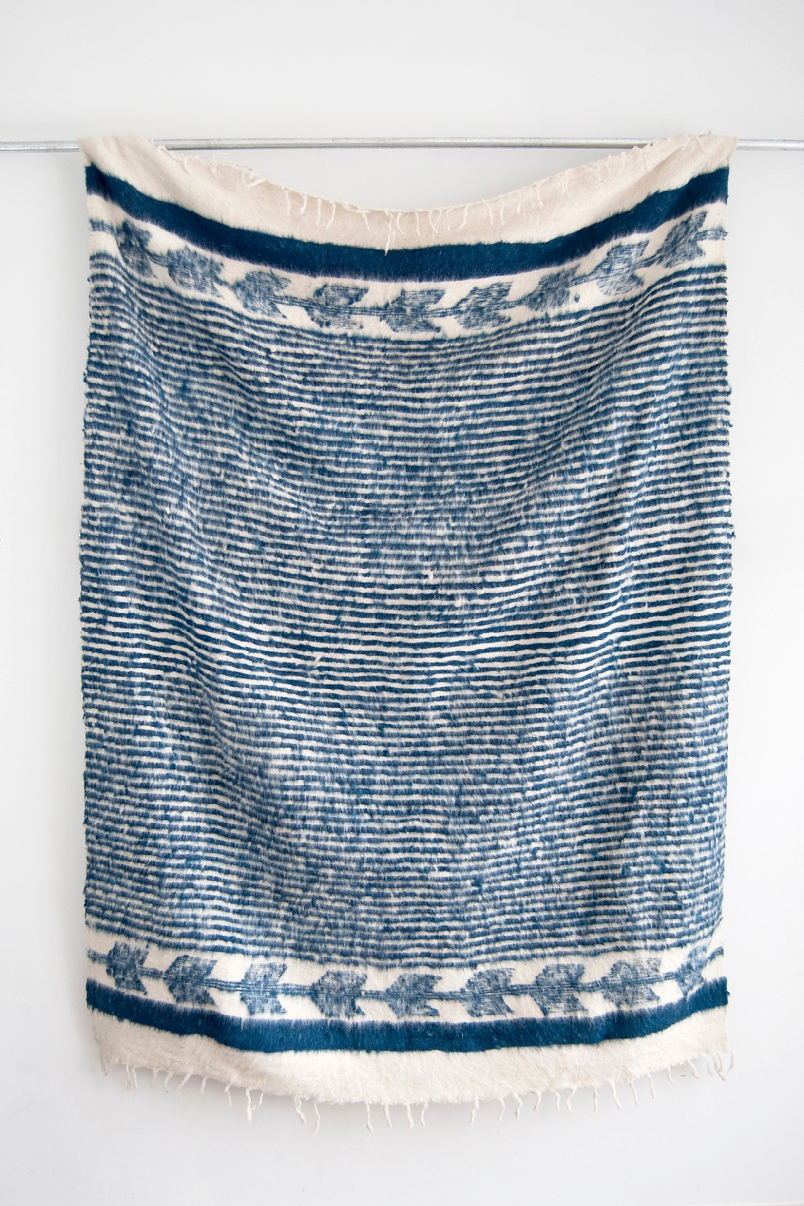 Queen sized ecru wool blanket with thin horizontal blue-indigo stripes throughout the body and blue-indigo arrow motif at both ends