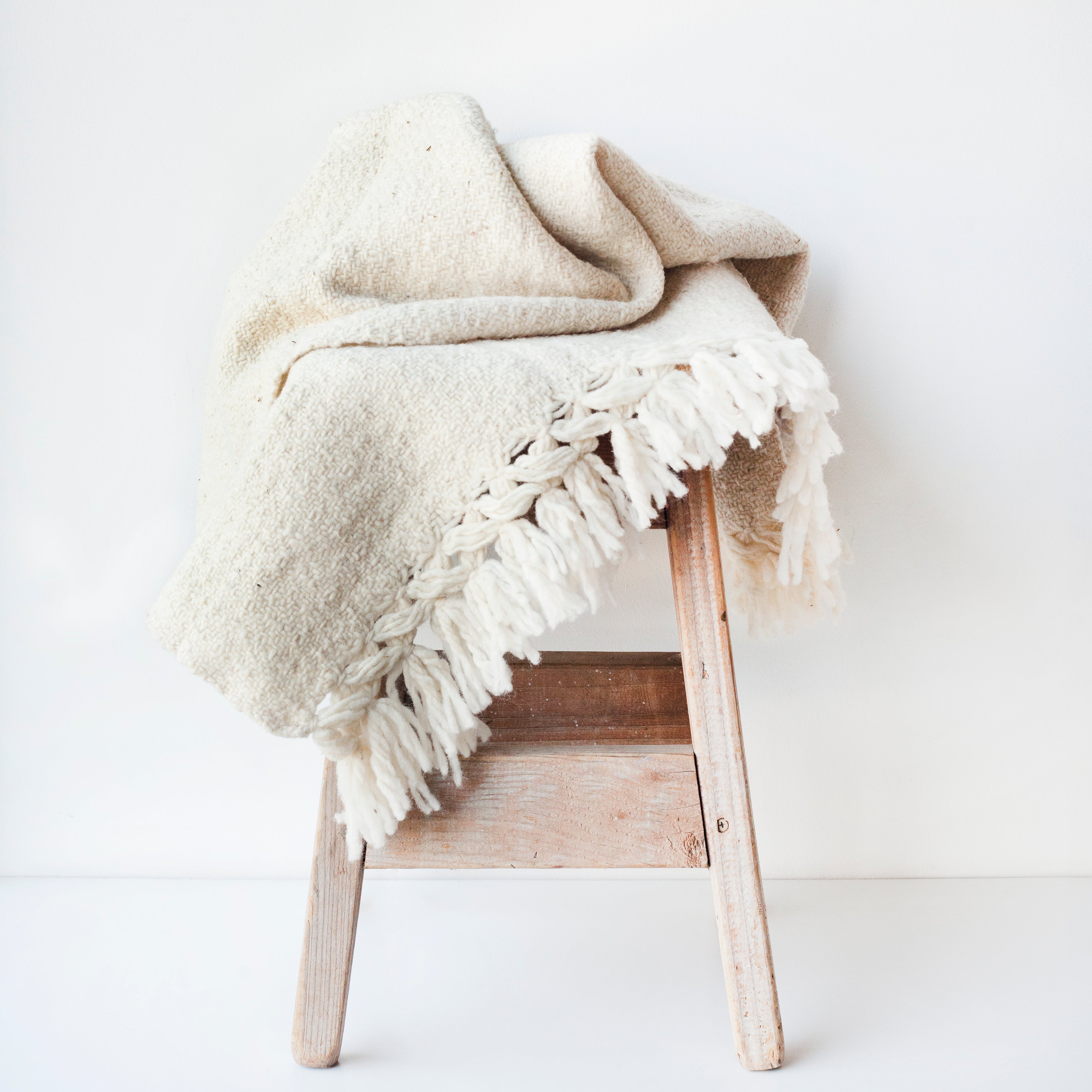 Cream wool throw blanket woven in a diamond pattern with hand tied tassels at each end draped over a wood stool