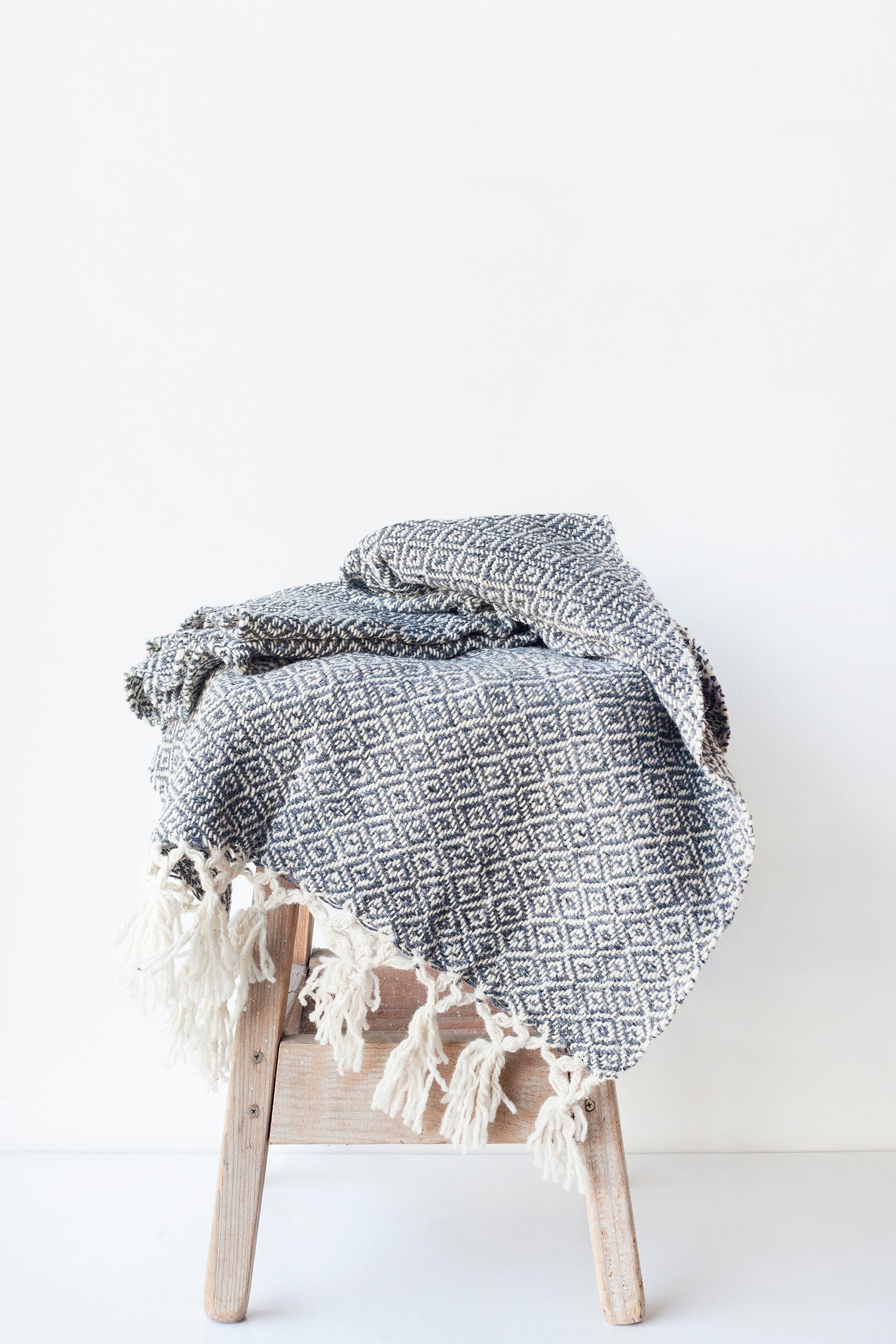 Charcoal grey and white wool throw blanket woven in a diamond pattern with tied tassels at each end draped over a wood stool