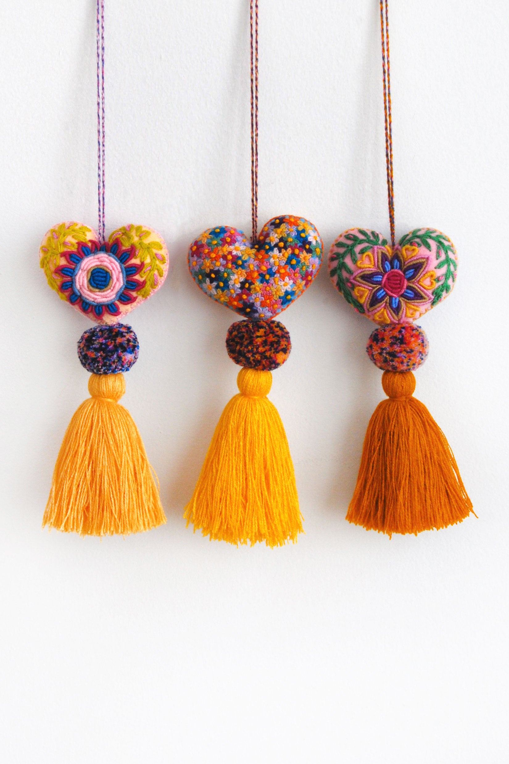 Three colorful plush felt hearts adorned with flower embroidery attached by multicolor speckled pom poms to tassels in varying shades of gold. Each heart is hanging from a string attached to the top of it.