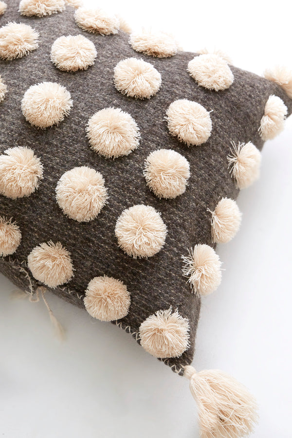 Detail of square woven grey wool throw pillow covered in cream pom poms with cream tassels in each corner