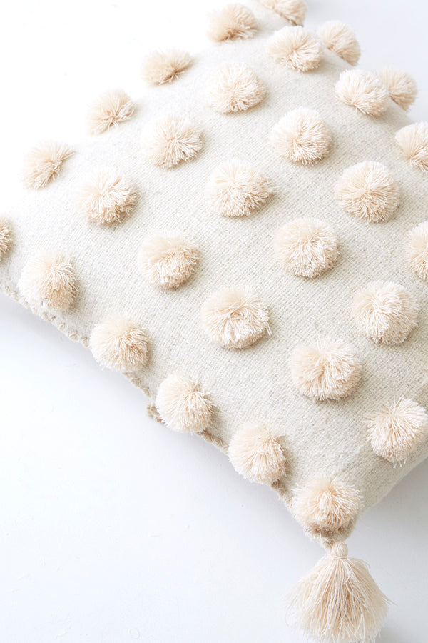 Detail of square woven wool cream throw pillow covered in cream pom poms with cream tassels in each corner