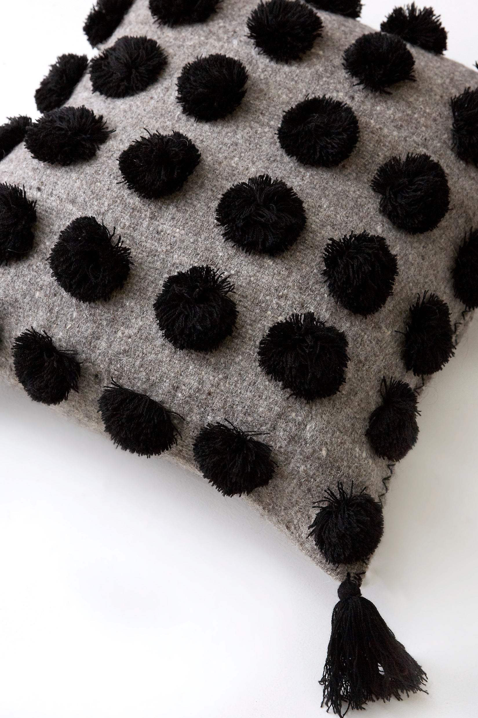 Detail of square woven wool grey throw pillow covered in black pom poms with black tassels in each corner