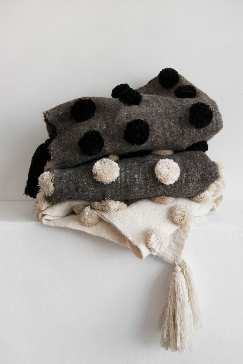 Three neutral-toned throws covered in pom poms stacked on top of each other