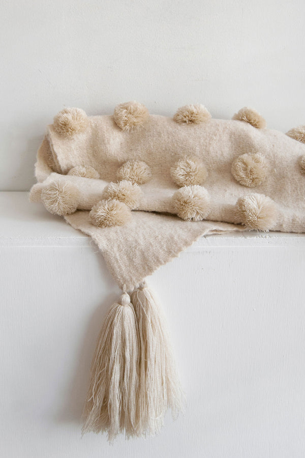 Detail of cream wool throw covered in rows of cream pom poms with cream tassels at each corner
