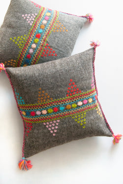 Two square woven grey wool accent pillows with colorful embroidery and multicolor fringe tassels at each corner