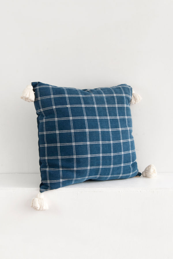 A square pillow with a white windowpane pattern over blue fabric sitting on a shelf. A short white tassel is attached to each corner of the pillow.