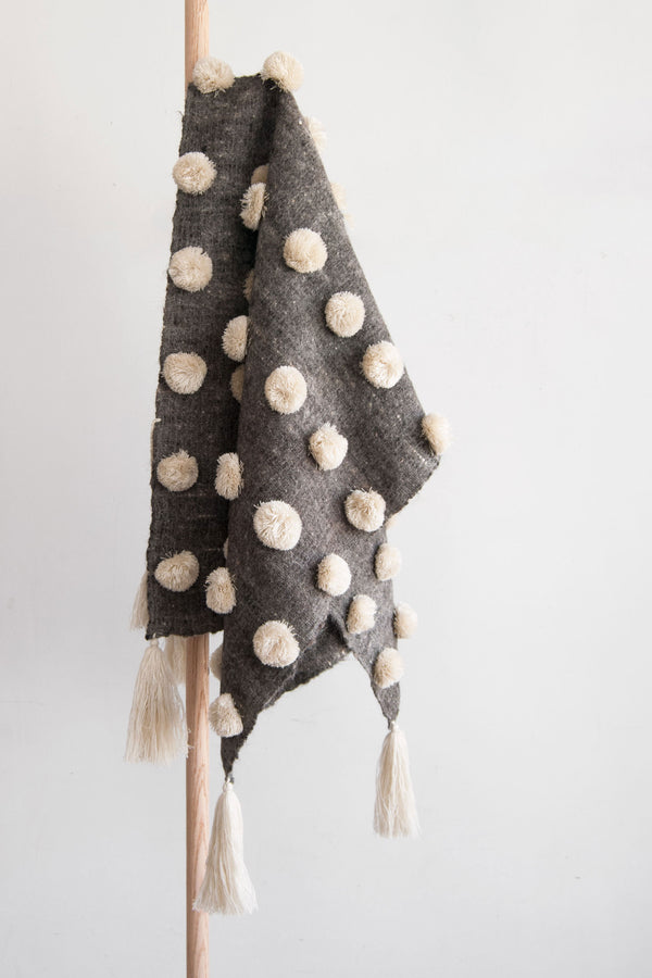 Grey wool throw covered in rows of cream pom poms with cream tassels at each corner