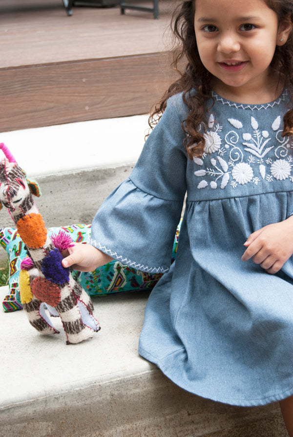 Four year old girl wearing Florcita dress looking at camera and smiling. She is holding a giraffe stuffed animal.