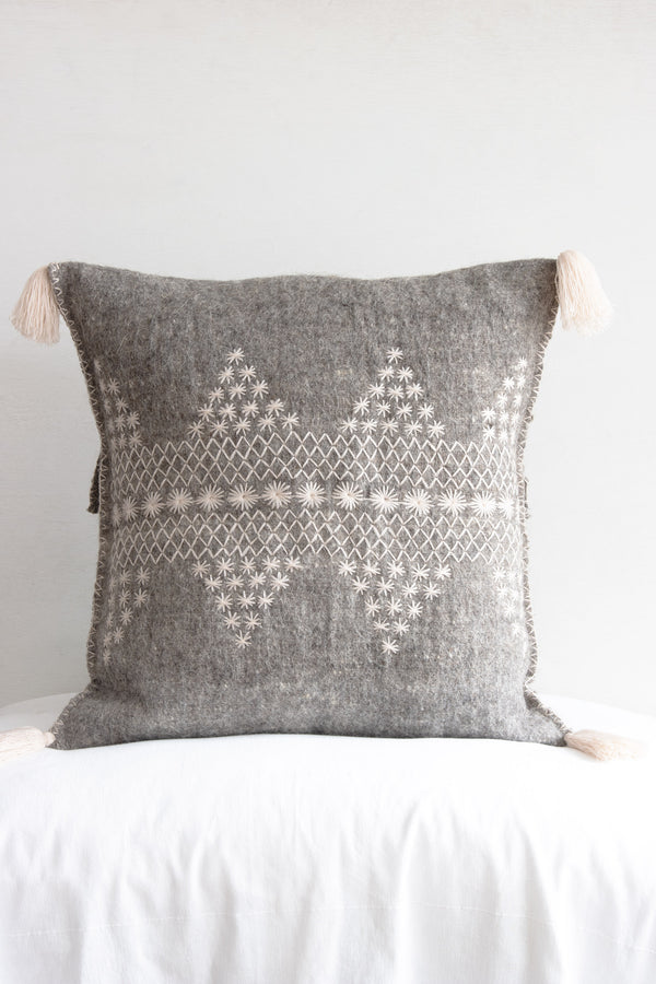 Square woven grey wool accent pillow with decorative cream embroidery and cream fringe tassels at each corner