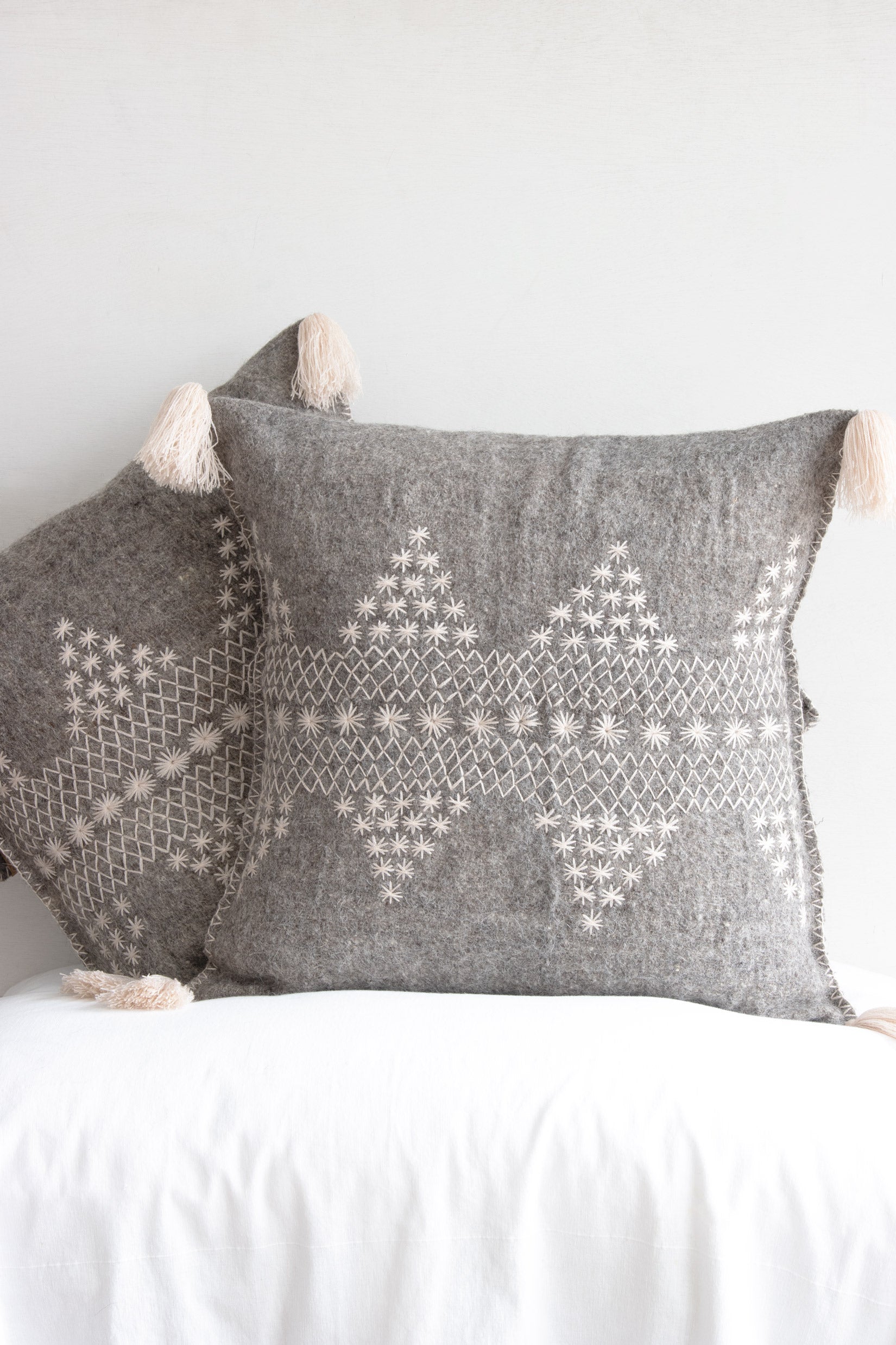 Two square woven grey wool accent pillows with decorative cream embroidery and cream fringe tassels at each corner