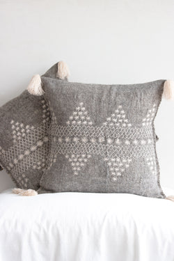 Two square woven grey wool accent pillows with decorative cream embroidery and cream fringe tassels at each corner