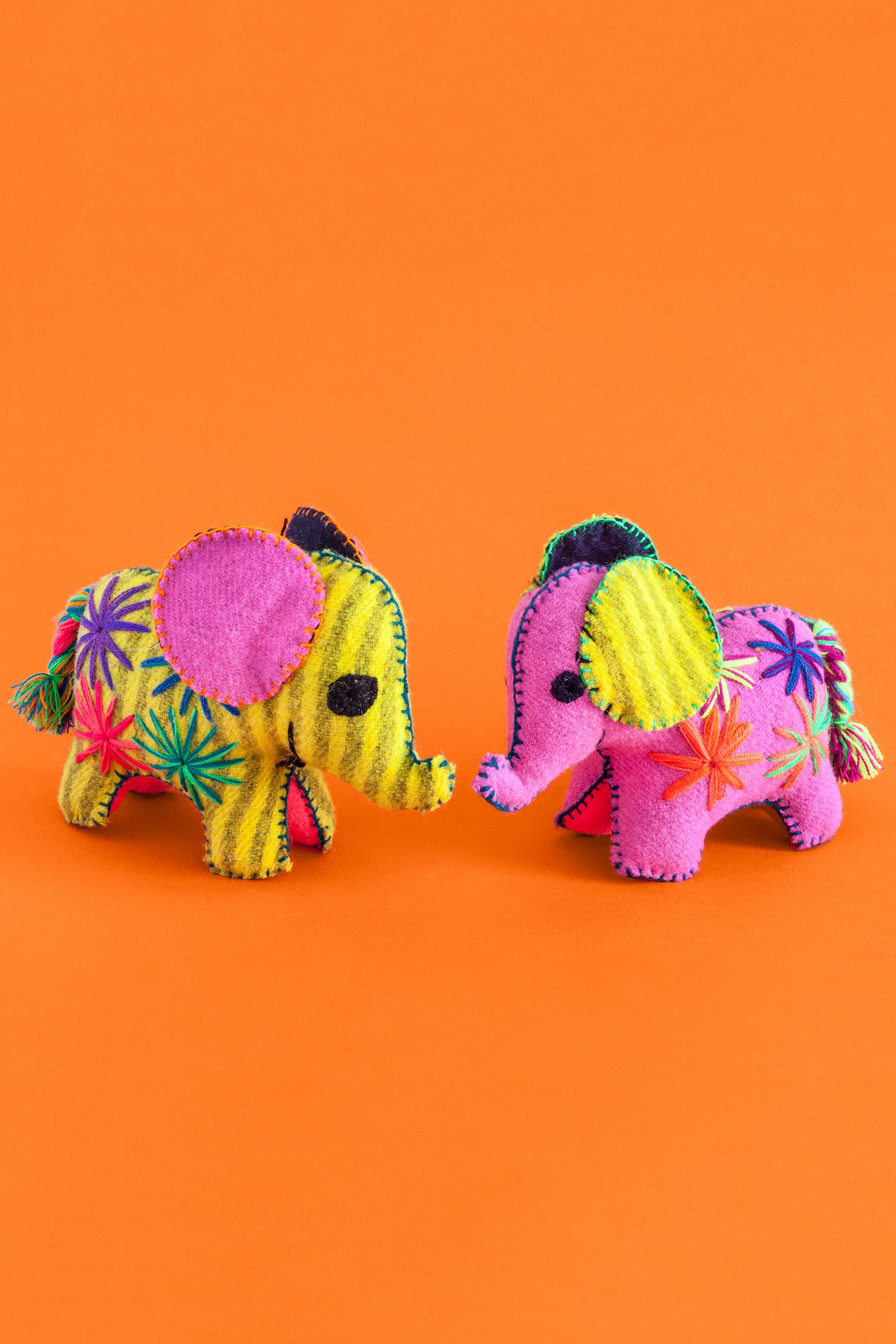 Two colorful felt elephant plush toys with a multicolor braided tails and hand-embroidered features and star designs.