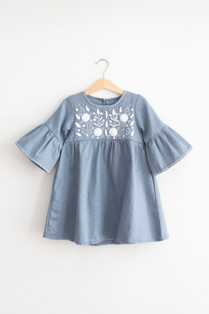 Kids light denim, knee-length dress with 3/4 bell sleeves. White, hand-embroidered floral pattern on chest with cross-stitch trim on sleeve ends and neckline