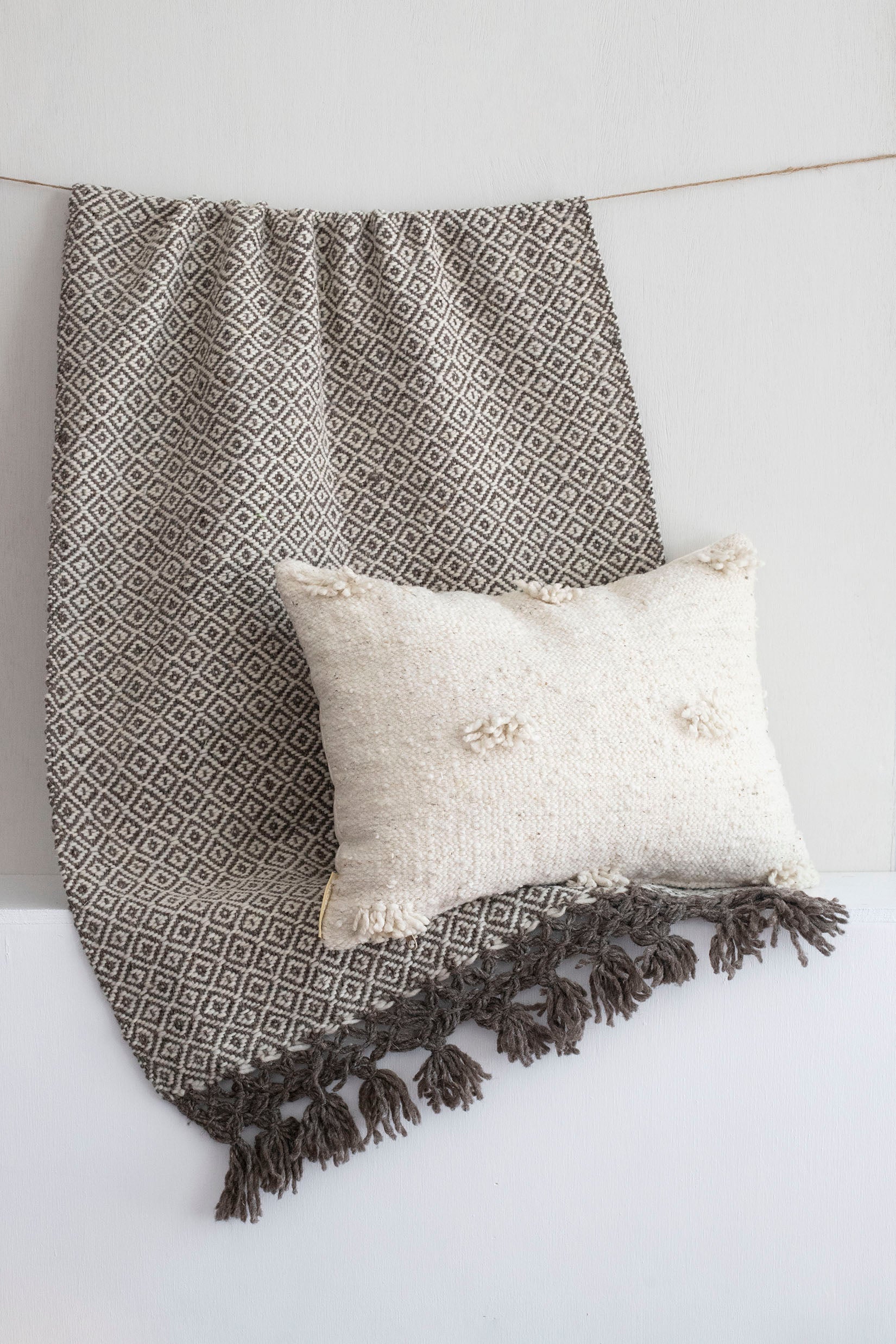 Small cream lumbar throw pillow with fringe accents sitting atop a dark brown and white wool throw in a diamond design.