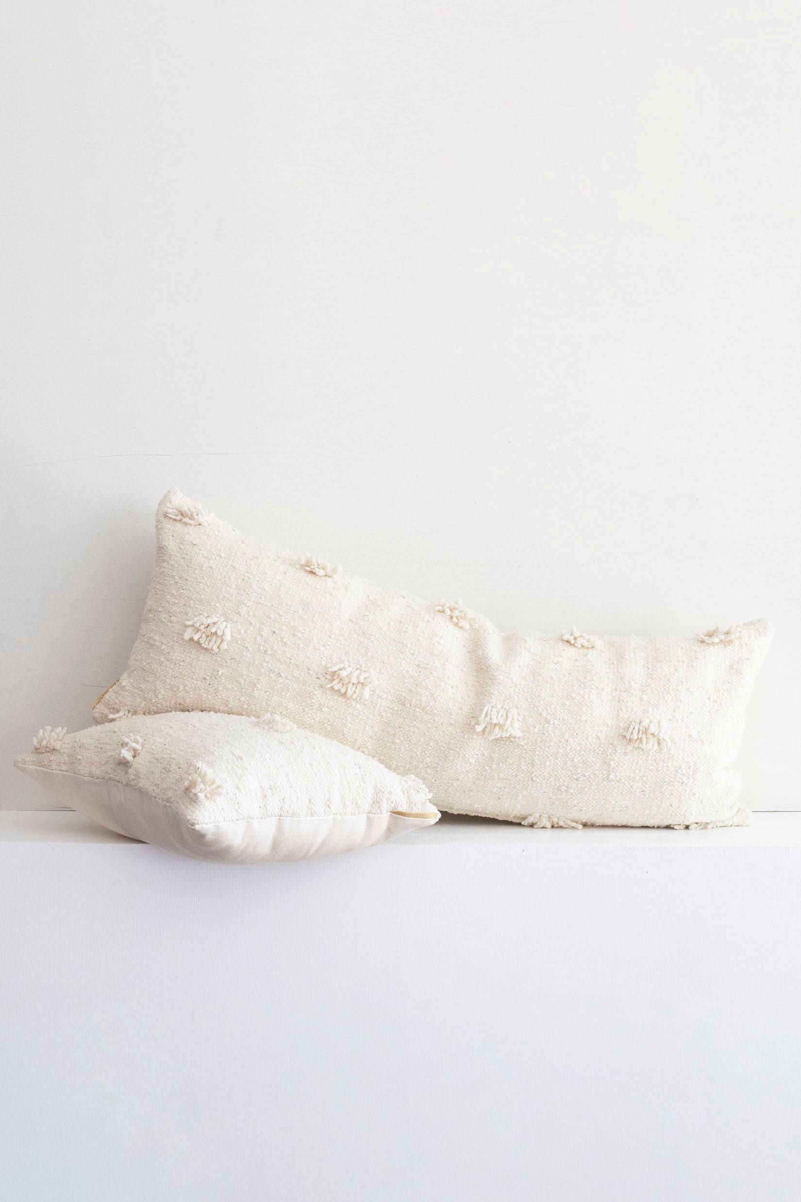 Large cream lumbar pillow with fringe accents laid on top of a smaller lumbar pillow in same style