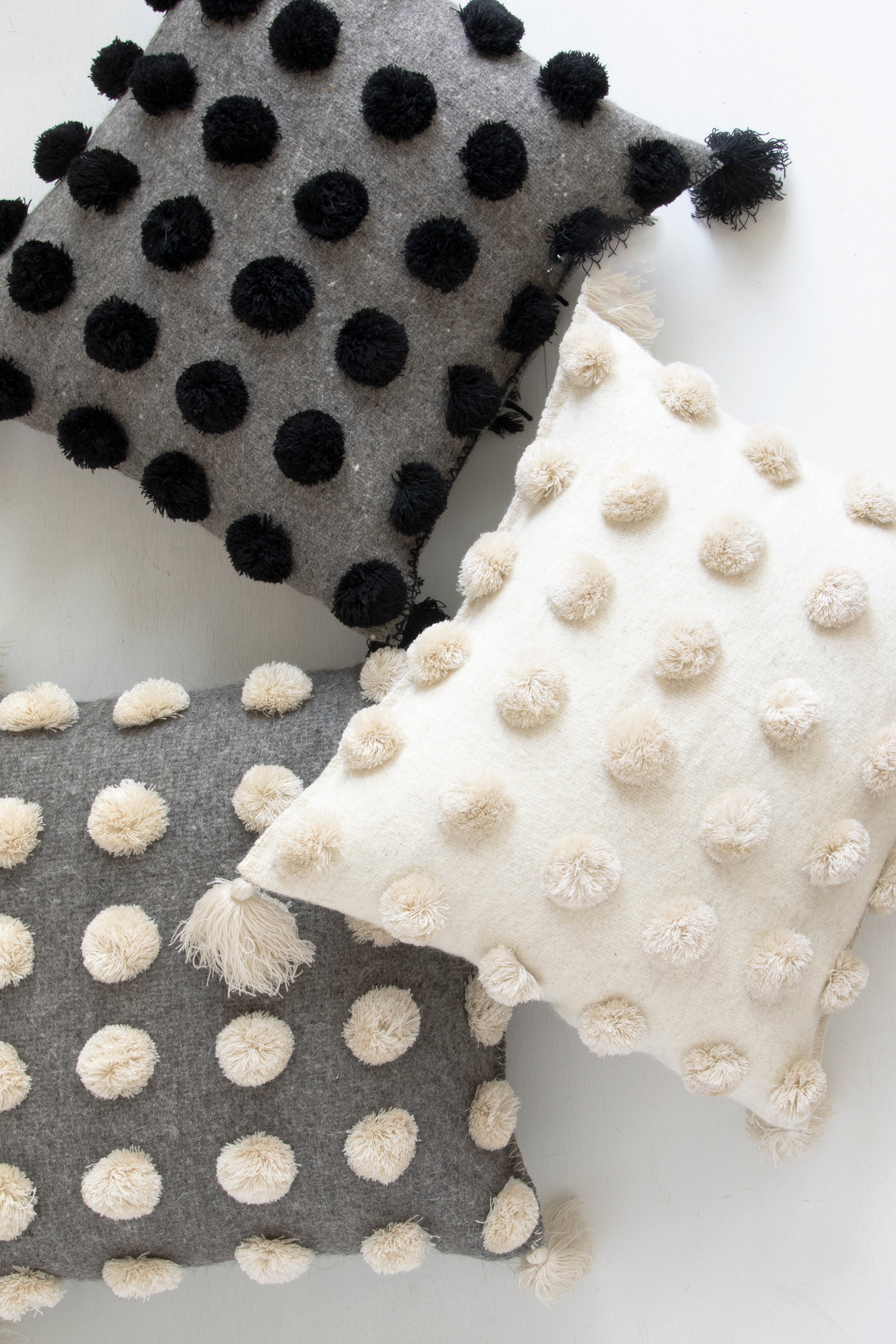 Three square woven wool throw pillows in different neutral colors covered in pom poms with tassels in each corner