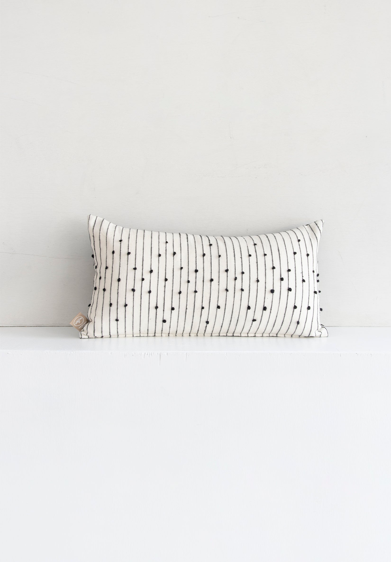 Lumbar white woven throw pillow with small black pom pom accents arranged in a repeating diamond pattern arranged along black lines stretching across the pillow.