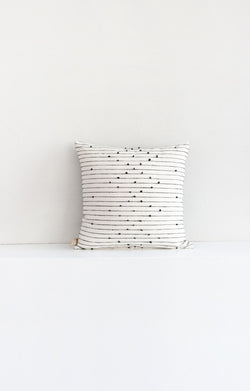 Square white woven throw pillow with small black pom pom accents arranged in a repeating diamond pattern arranged along black lines stretching across the pillow.