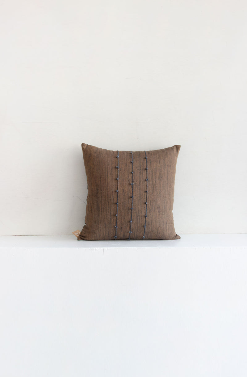Square brown woven throw pillow with small grey pom poms arranged along three vertical grey lines stretching across the middle of the pillow.