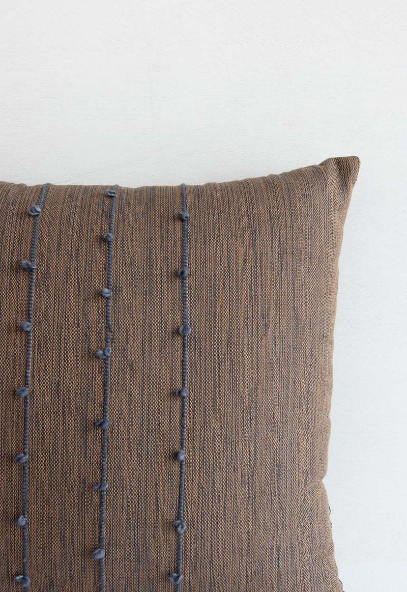 Detail of brown and grey woven textile of pillow, showing the three vertical grey lines and small grey pom poms arranged along them.