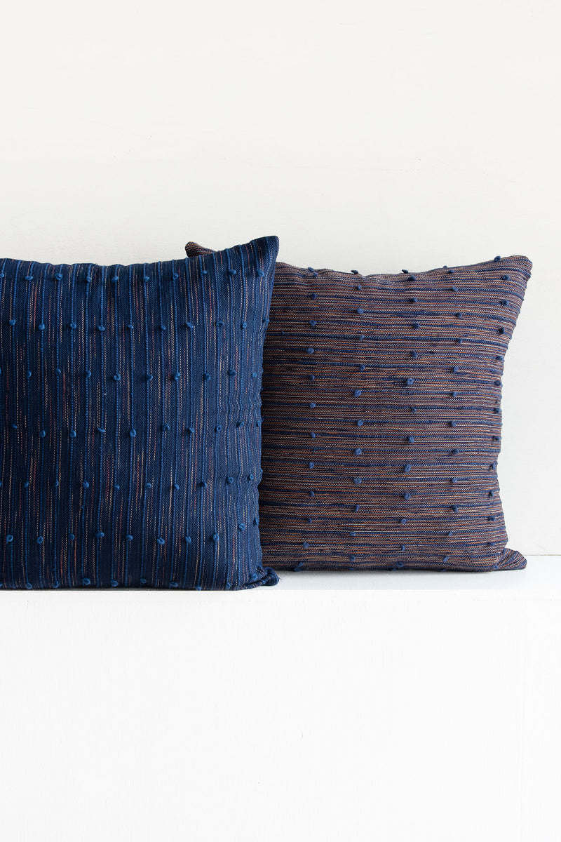 Two complementary pillows sitting side-by-side, one a mix of coral red and navy and the other navy with beige and coral red accents throughout. Both have raised navy lines running across them, with small navy pom poms arranged along these lines.