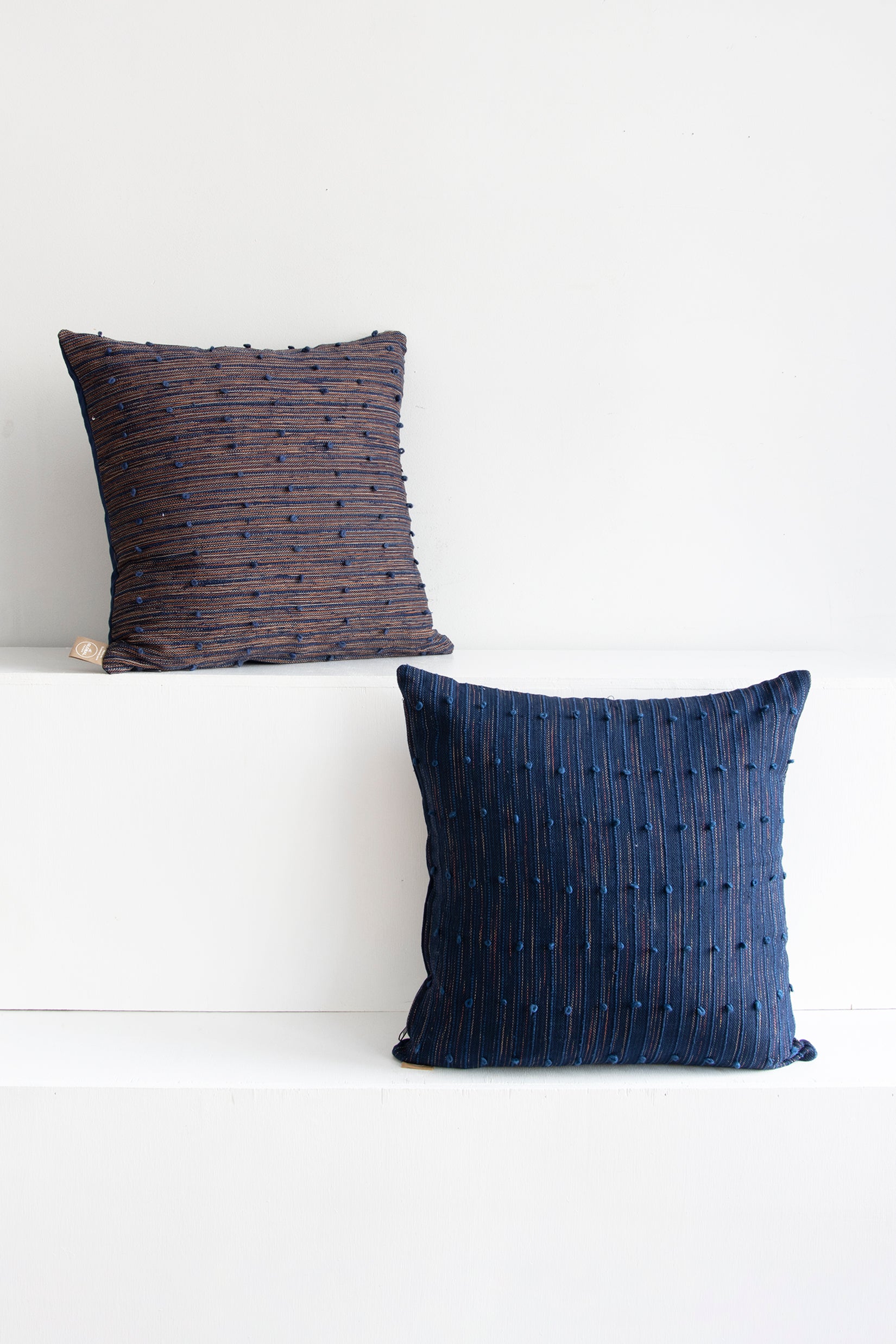 Two complementary pillows, one a mix of coral red and navy and the other navy with beige and coral red accents throughout. Both have raised navy lines running across them, with small navy pom poms arranged along these lines.