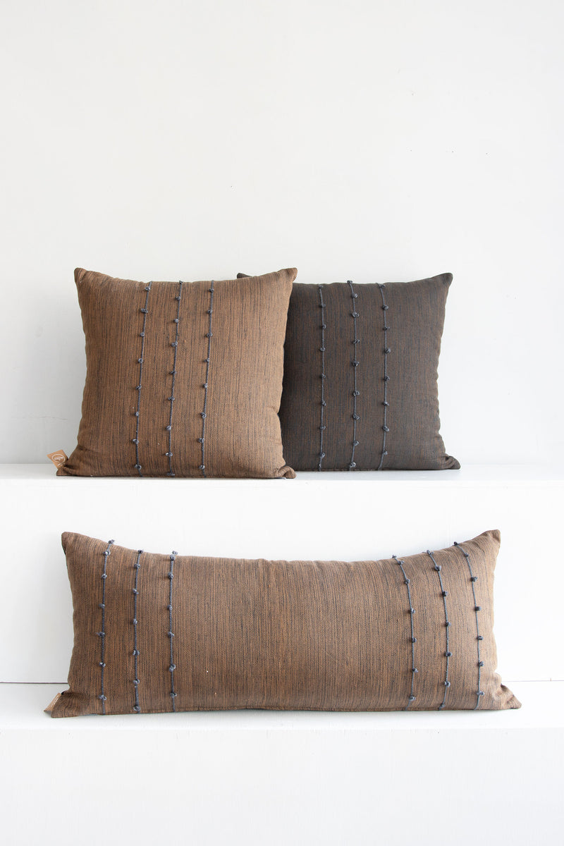 Two complementary woven square throw pillows side-by-side, one brown and one dark brown, and a large brown lumbar pillow sitting below them. All the pillows have small grey pom poms arranged along three vertical grey lines stretching across the pillows.