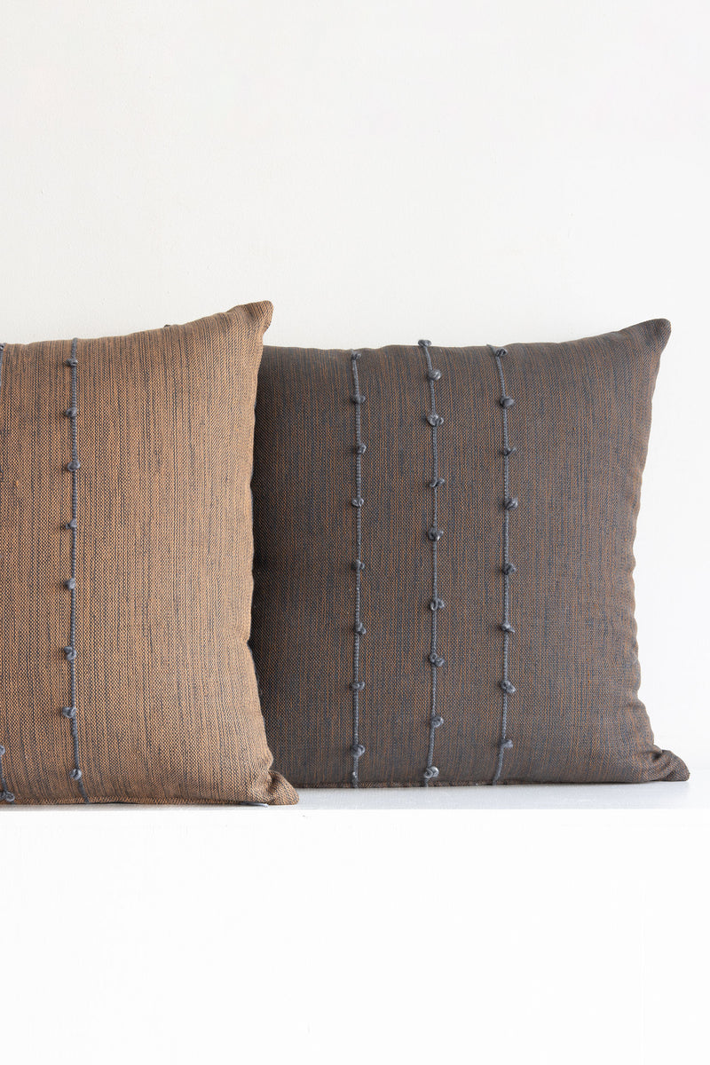 Two complementary woven square throw pillows side-by-side, one brown and one dark brown, both with small grey pom poms arranged along three vertical grey lines stretching across the middle of the pillows.