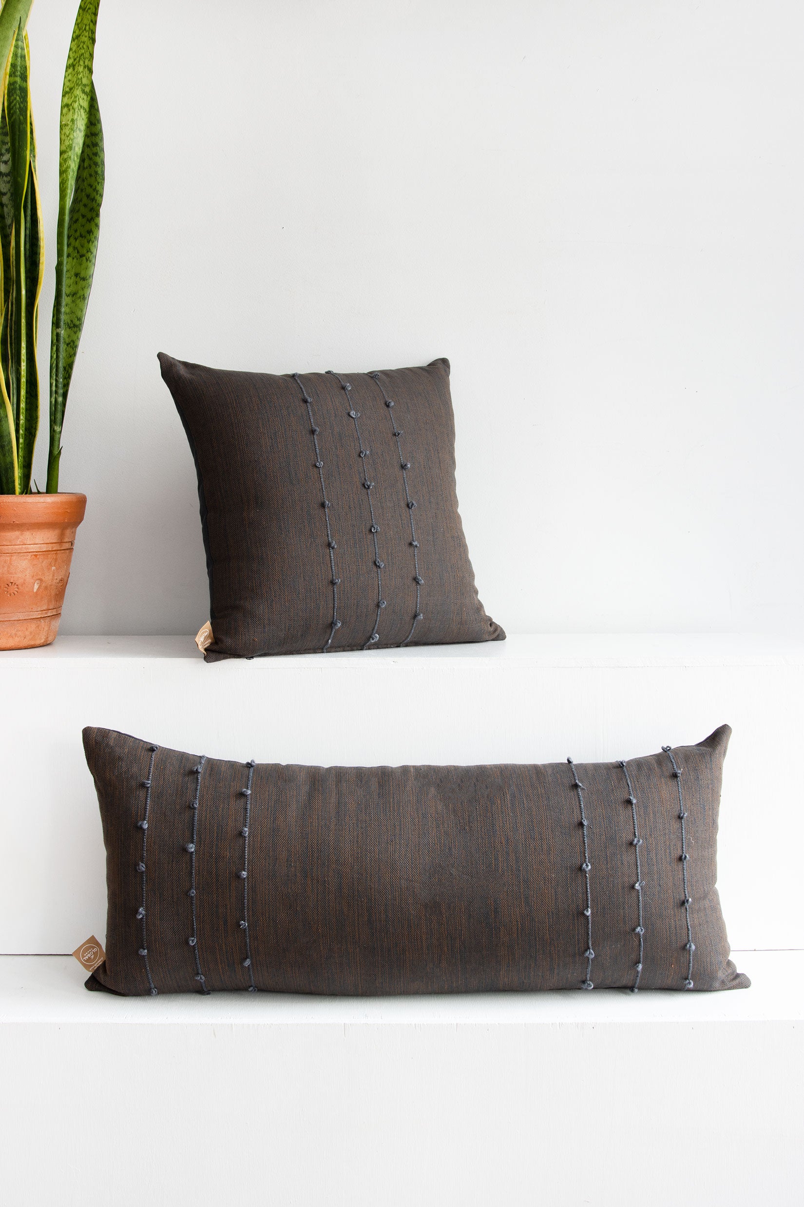 Two dark brown tight woven pillows in different shapes with small grey pom poms arranged along three vertical grey lines stretching across the pillows.