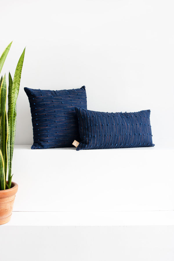Two woven navy pillows in different shapes with accents of beige and coral red and small navy pom poms arranged along raised navy lines spread across the pillows.