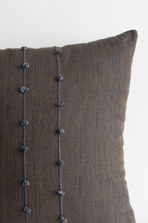 Detail of grey and brown woven textile of pillow, showing the vertical grey lines and small grey pom poms arranged along them.