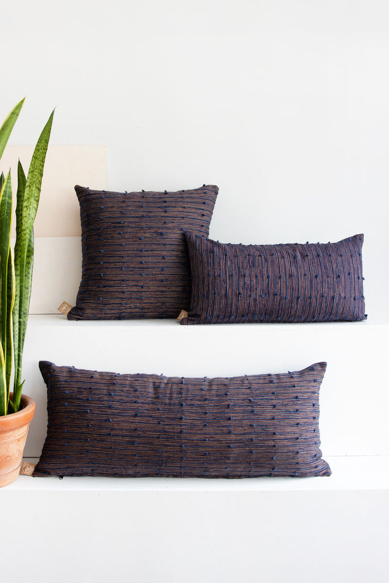Three woven pillows in different shapes and sizes that look like a mix between navy and coral red. All three have raised navy lines running across them, with small navy pom poms arranged along these lines.
