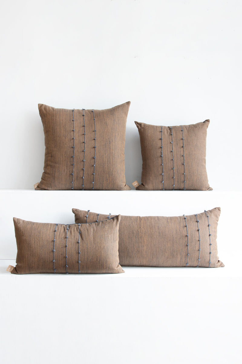 Four tight-woven brown pillows in different shapes and sizes with small grey pom poms arranged along three vertical grey lines stretching across the pillows.