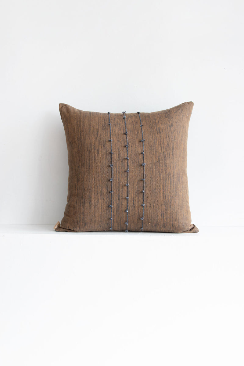 Large square brown woven throw pillow with small grey pom poms arranged along three vertical grey lines stretching across the middle of the pillow.