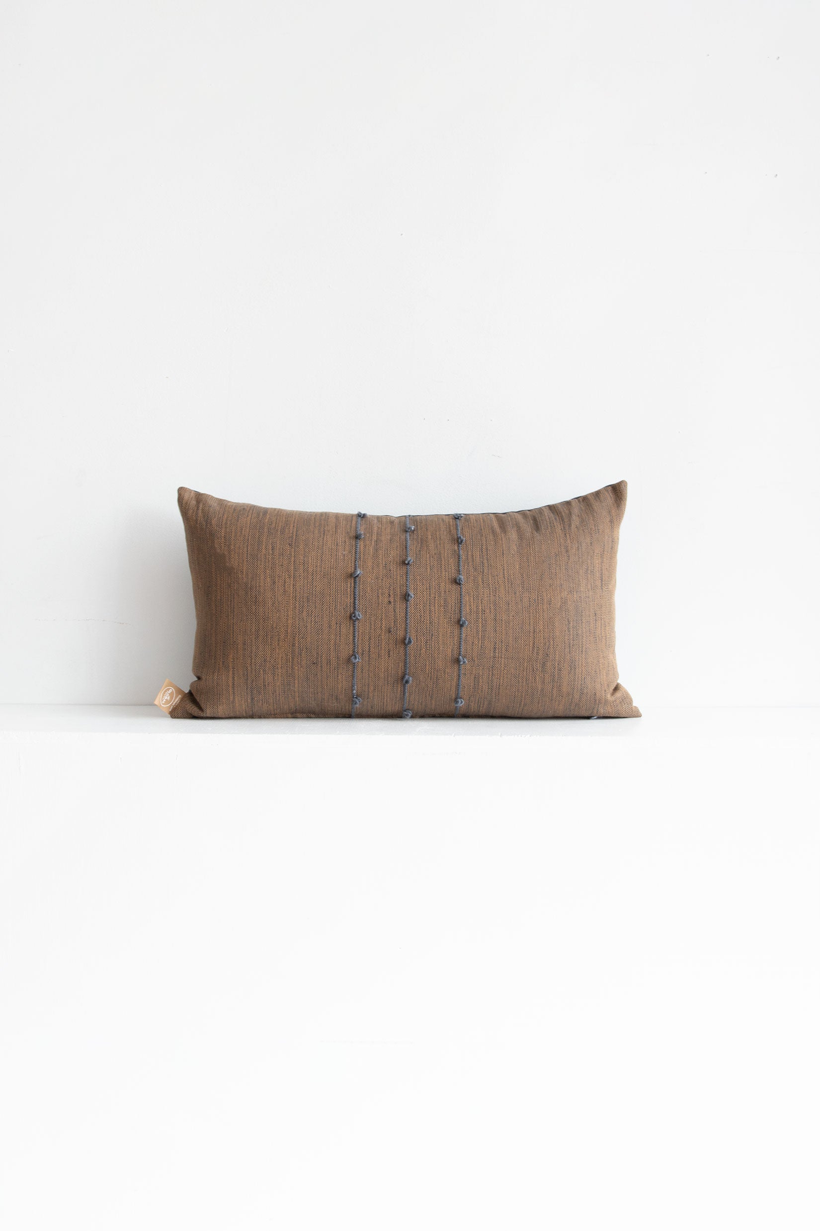 Lumbar brown woven throw pillow with small grey pom poms arranged along three vertical grey lines stretching across the middle of the pillow.