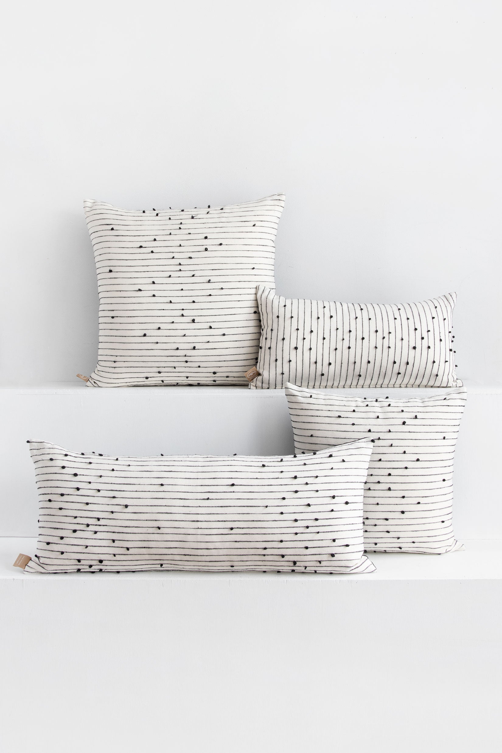 Four tight-woven white pillows in different shapes and sizes with small black pom poms arranged in a repeating diamond pattern arranged along black lines stretching across the pillows.