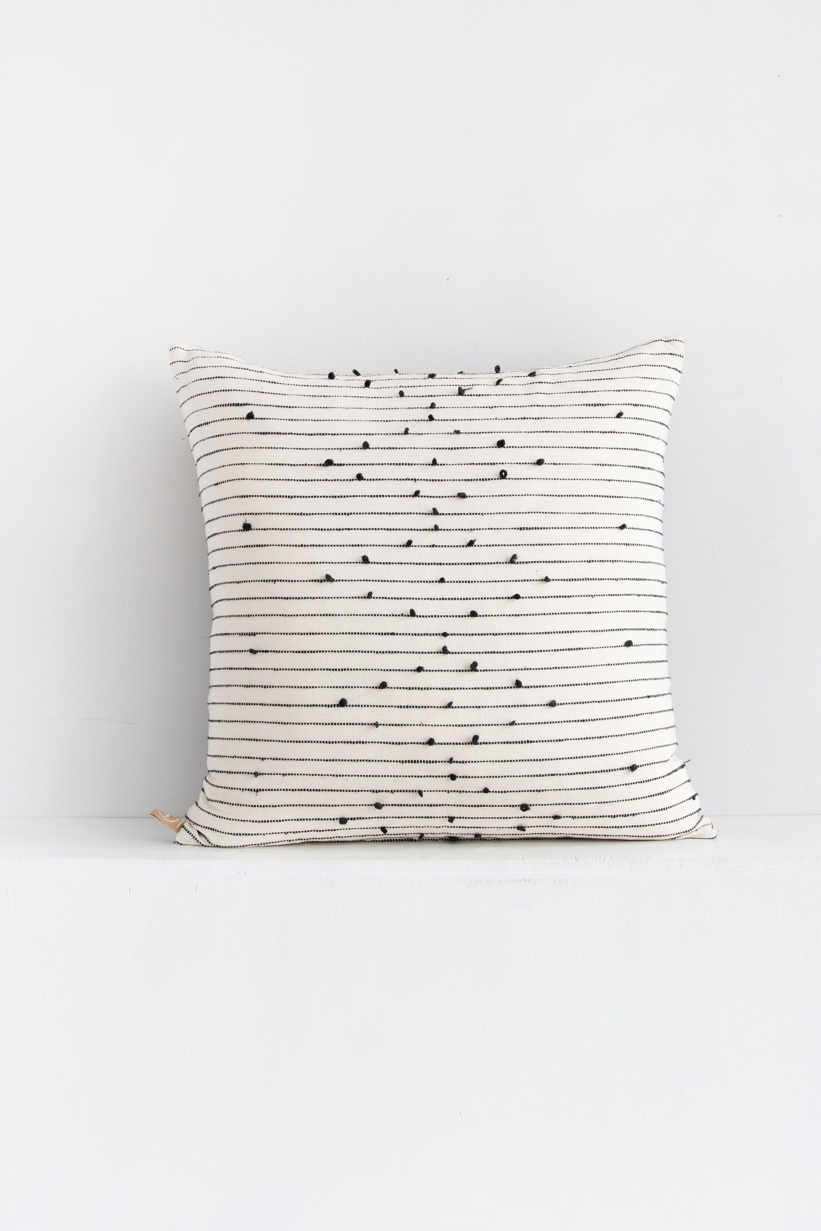 Large square white woven throw pillow with small black pom pom accents arranged in a repeating diamond pattern arranged along black lines stretching across the pillow.
