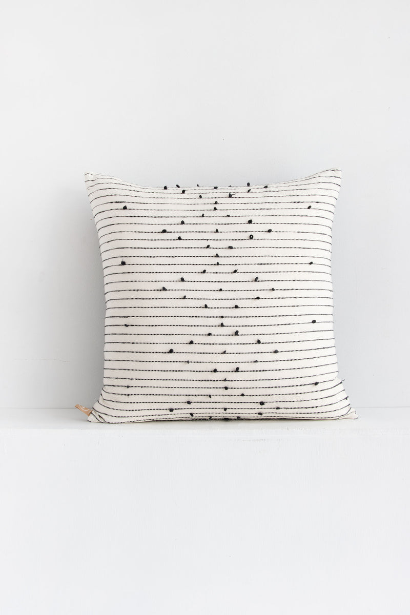 Large square white woven throw pillow with small black pom pom accents arranged in a repeating diamond pattern arranged along black lines stretching across the pillow.