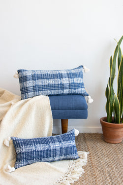 Two blue and white pattern textile lumbar pillows with white tassels at each corner in two sizes sitting on a couch and a blanket.