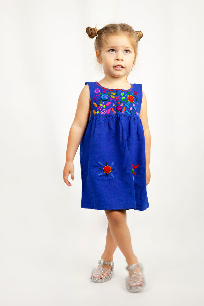 Child wearing a sleeveless royal blue knee-length sun dress. There are multicolor hand embroidered floral elements on the chest and skirt of the dress and cross-stitch trim around the neckline.