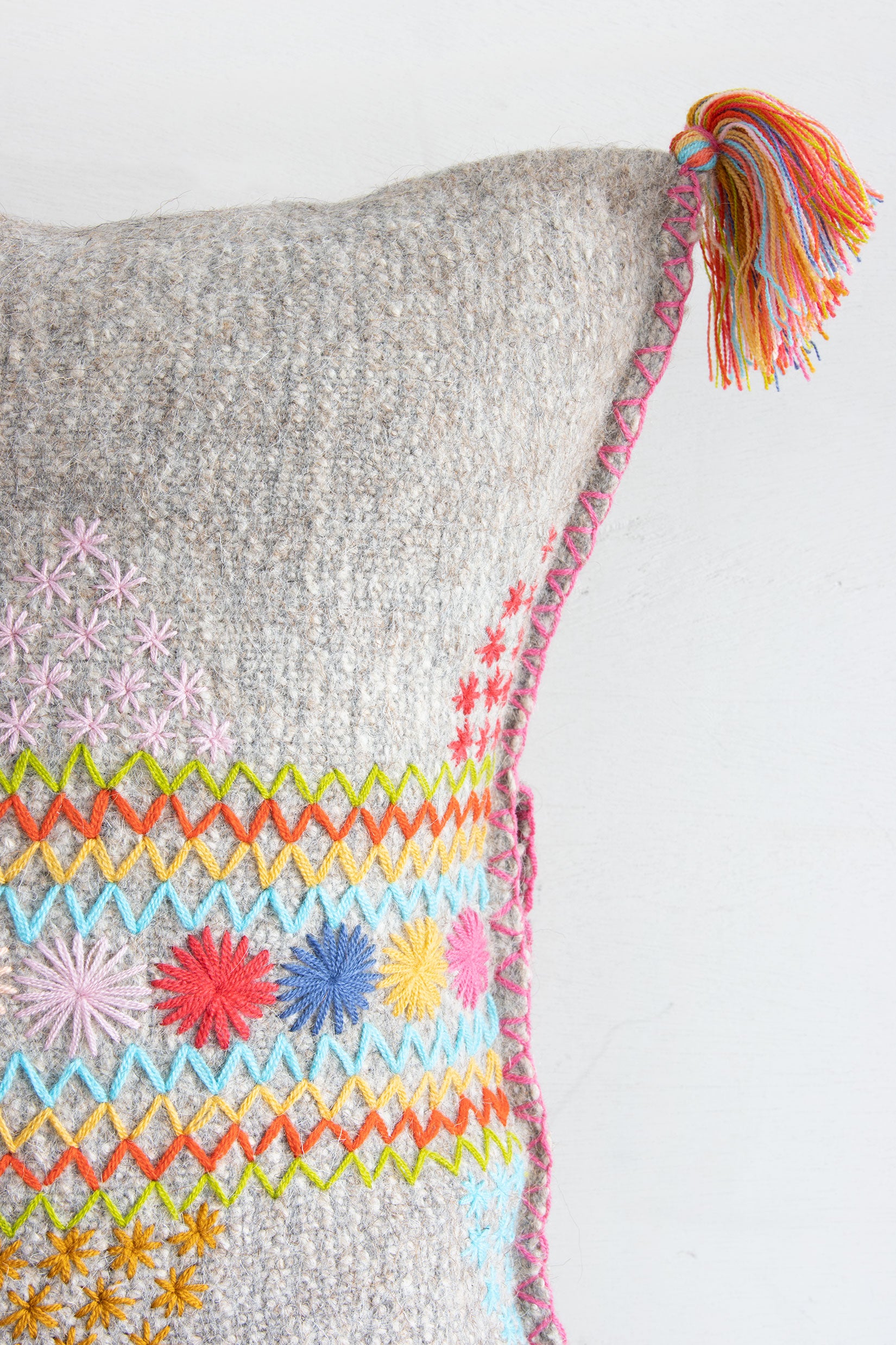 Detail of woven light grey wool throw pillow showing multicolor star and zig-zag embroidery, corner tassel, and hand-stitched seam.