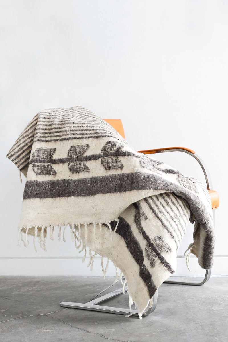 Queen sized ecru wool blanket with horizontal thin grey stripes throughout the body and grey arrow motif at both ends draped over a chair