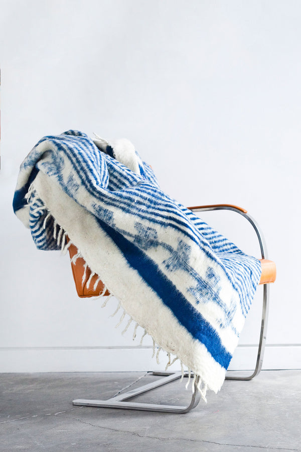 Queen sized ecru wool blanket with horizontal thin blue indigo stripes throughout the body and blue-indigo arrow motif at both ends draped over a chair