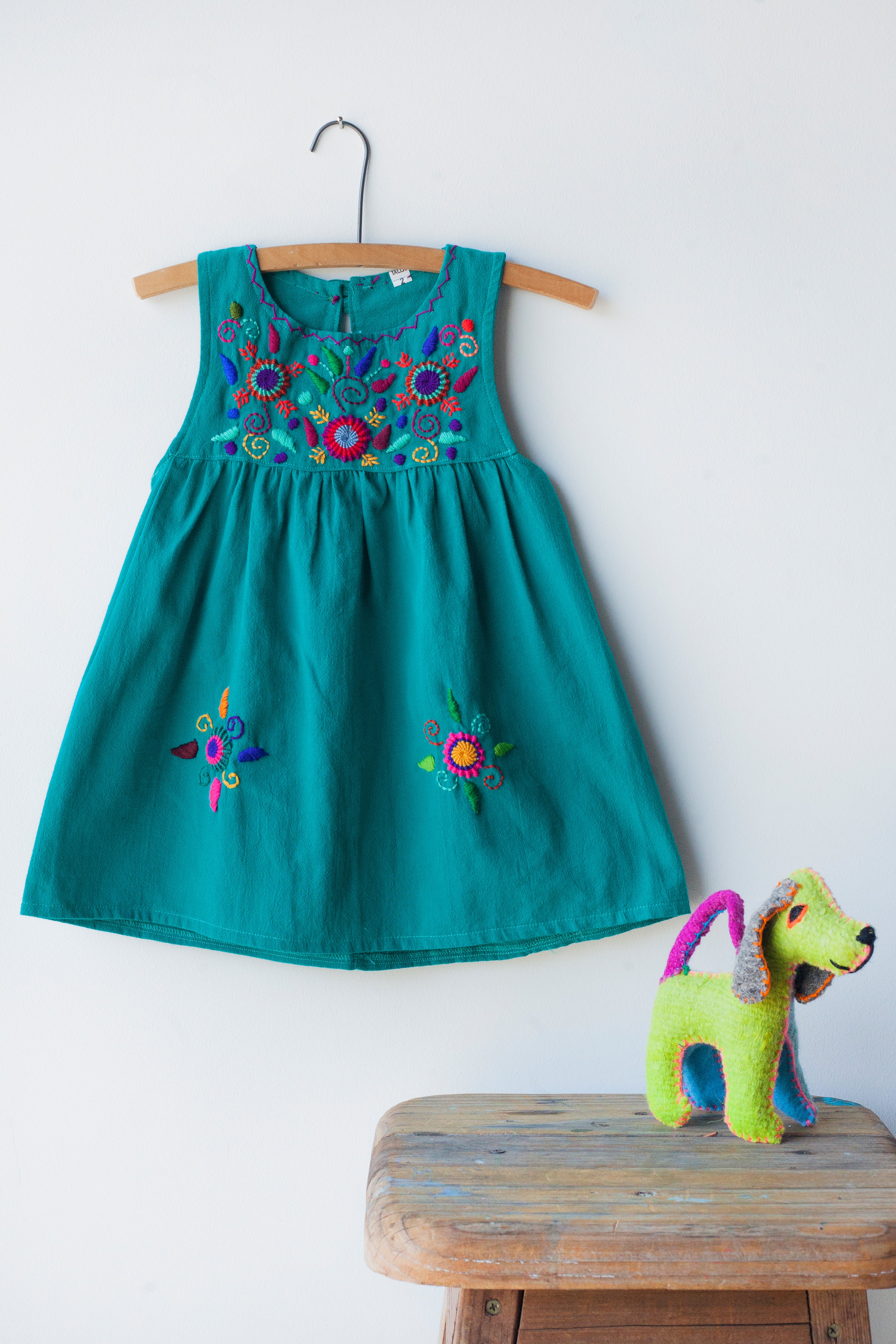 Kids sleeveless teal sun dress hanging with multicolor hand-embroidered floral pattern on chest and two hand-embroidered flowers near the bottom of the skirt on the front left and right.