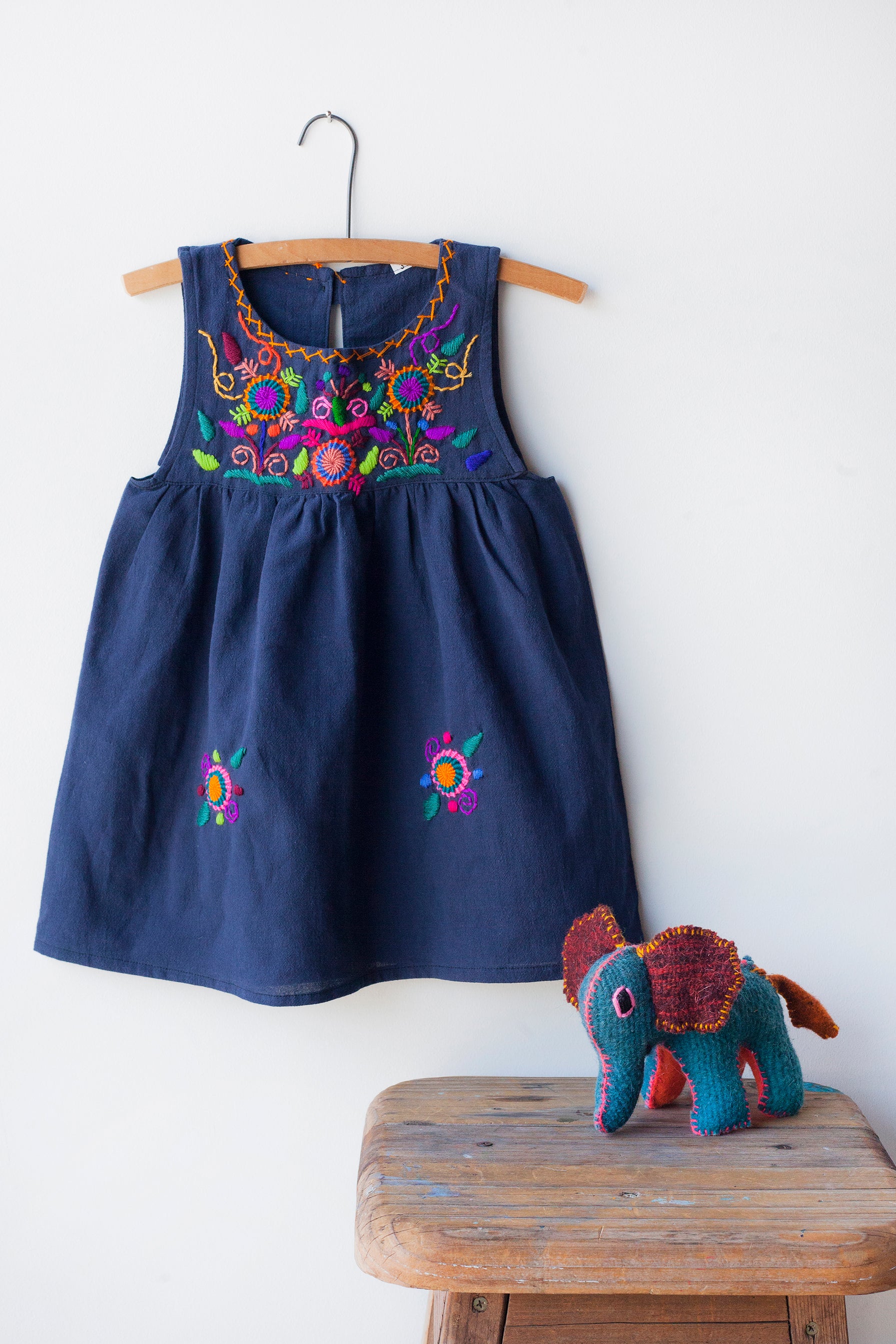 Kids sleeveless navy sun dress hanging with multicolor hand-embroidered floral pattern on chest and two hand-embroidered flowers near the bottom of the skirt on the front left and right.