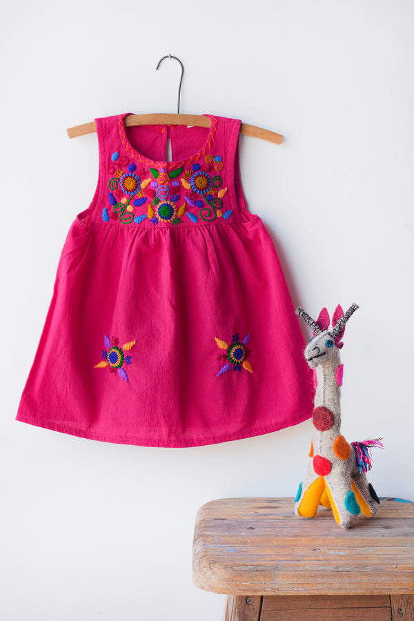 Kids sleeveless magenta sun dress hanging with multicolor hand-embroidered floral pattern on chest and two hand-embroidered flowers near the bottom of the skirt on the front left and right.