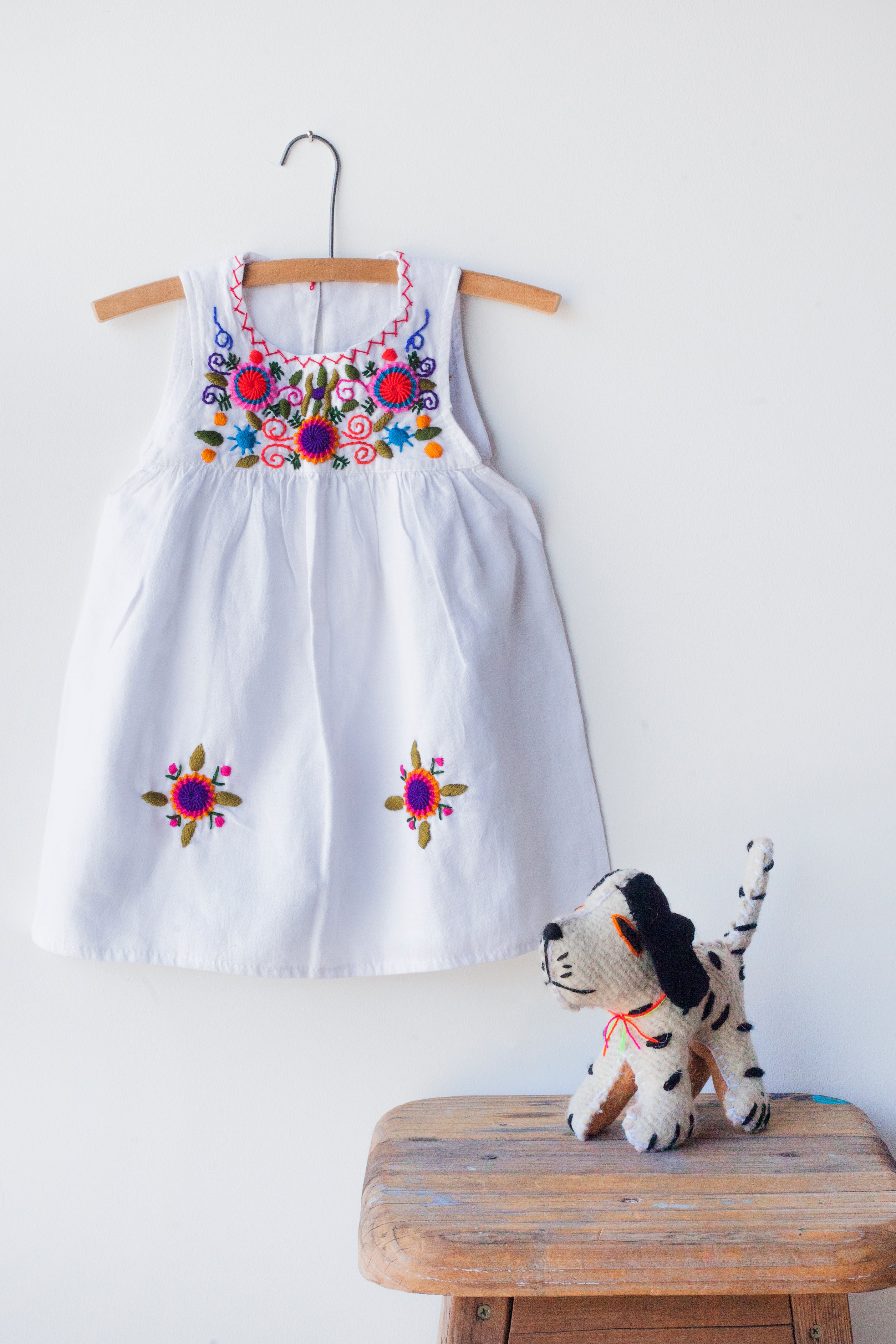 Kids sleeveless white sun dress hanging with multicolor hand-embroidered floral pattern on chest and two hand-embroidered flowers near the bottom of the skirt on the front left and right.