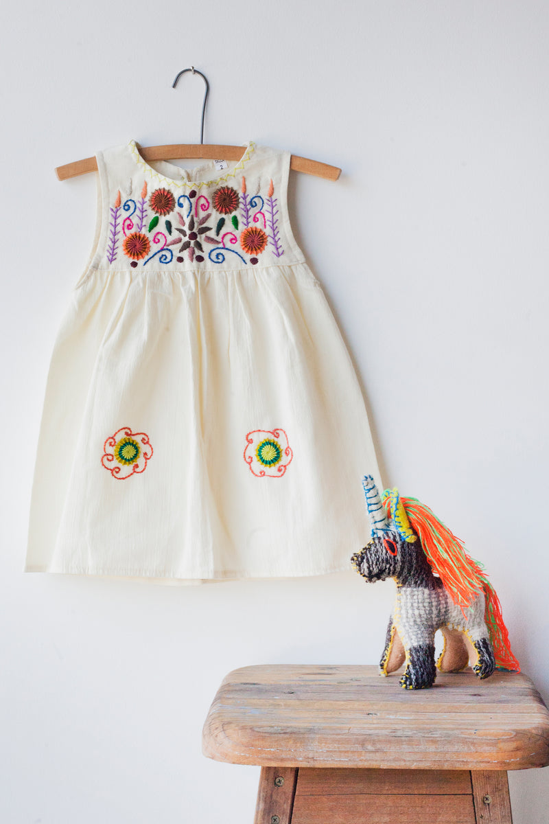 Kids sleeveless cream sun dress hanging with multicolor hand-embroidered floral pattern on chest and two hand-embroidered flowers near the bottom of the skirt on the front left and right.